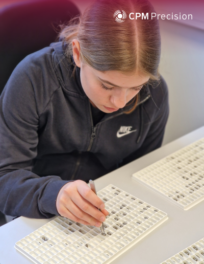 A girl works with focused attention at a table, sorting small metal parts into a plastic tray using tweezers.