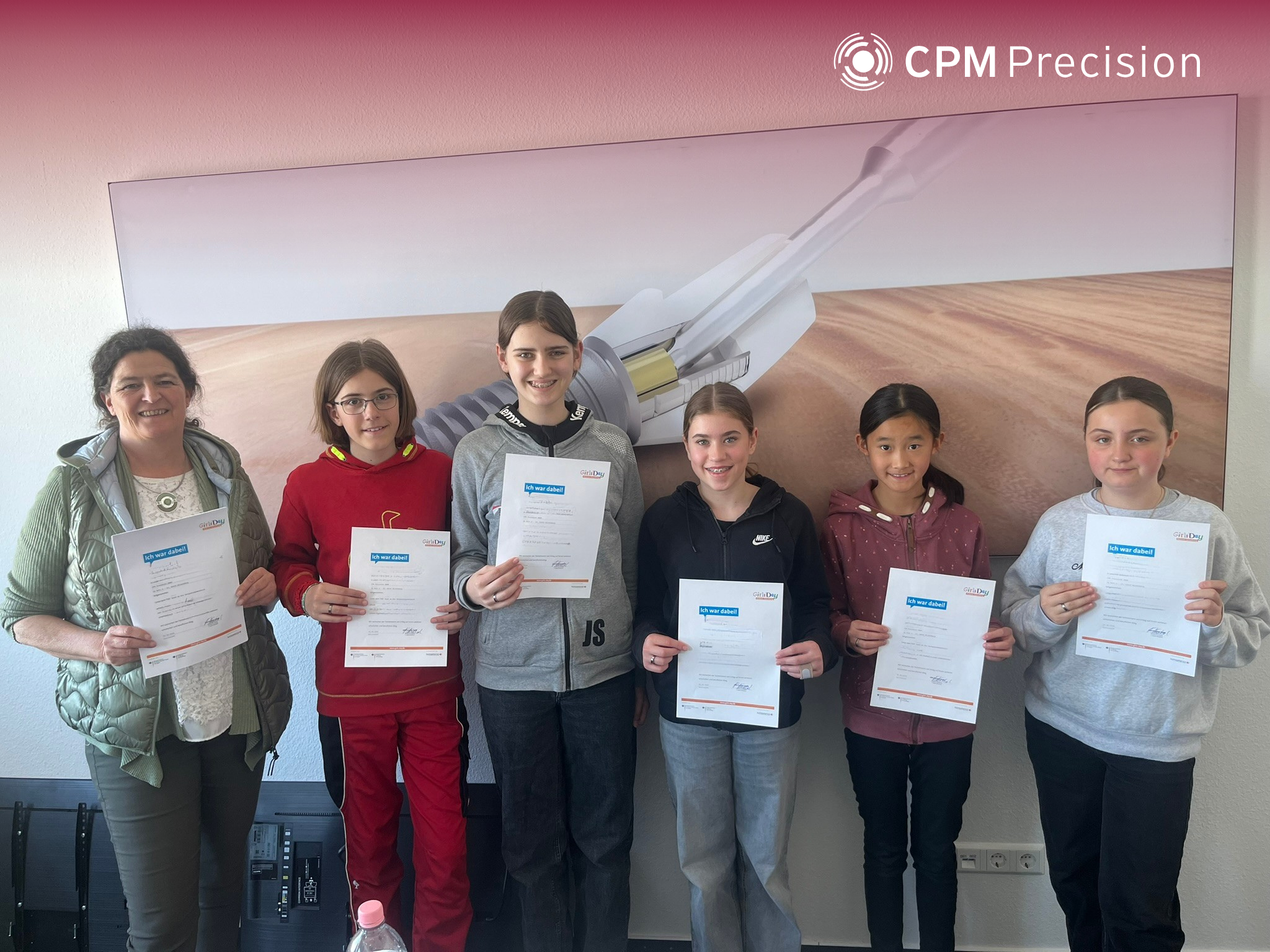 Group photo of the Girls’ Day participants at CPM Precision: Five girls and one adult supervisor stand side by side in front of a large image of a technical precision component. All are holding participation certificates and looking at the camera.