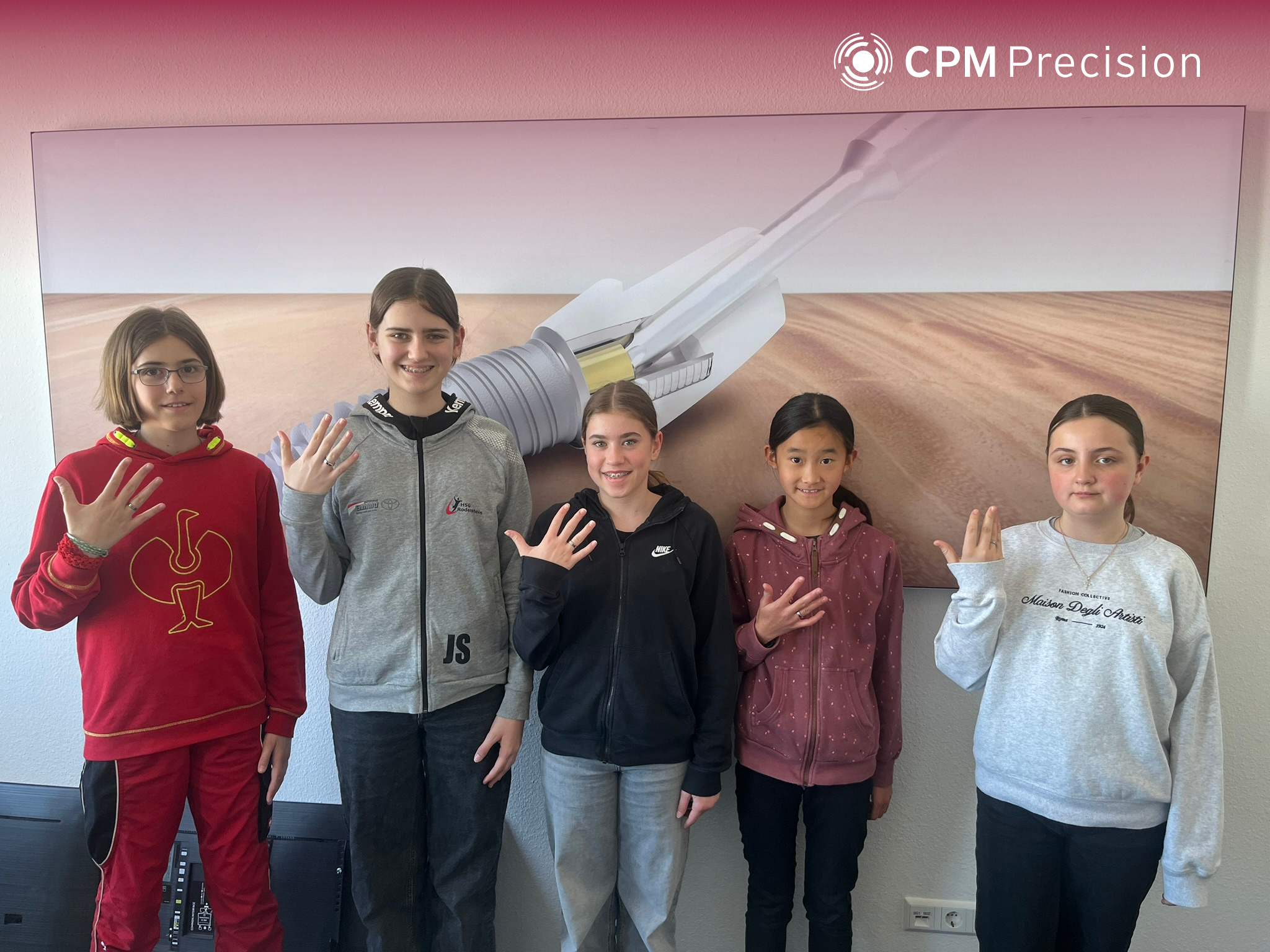 Five Girls’ Day participants stand side by side in front of a large image of a technical precision component, showing the titanium rings they made themselves on their hands.